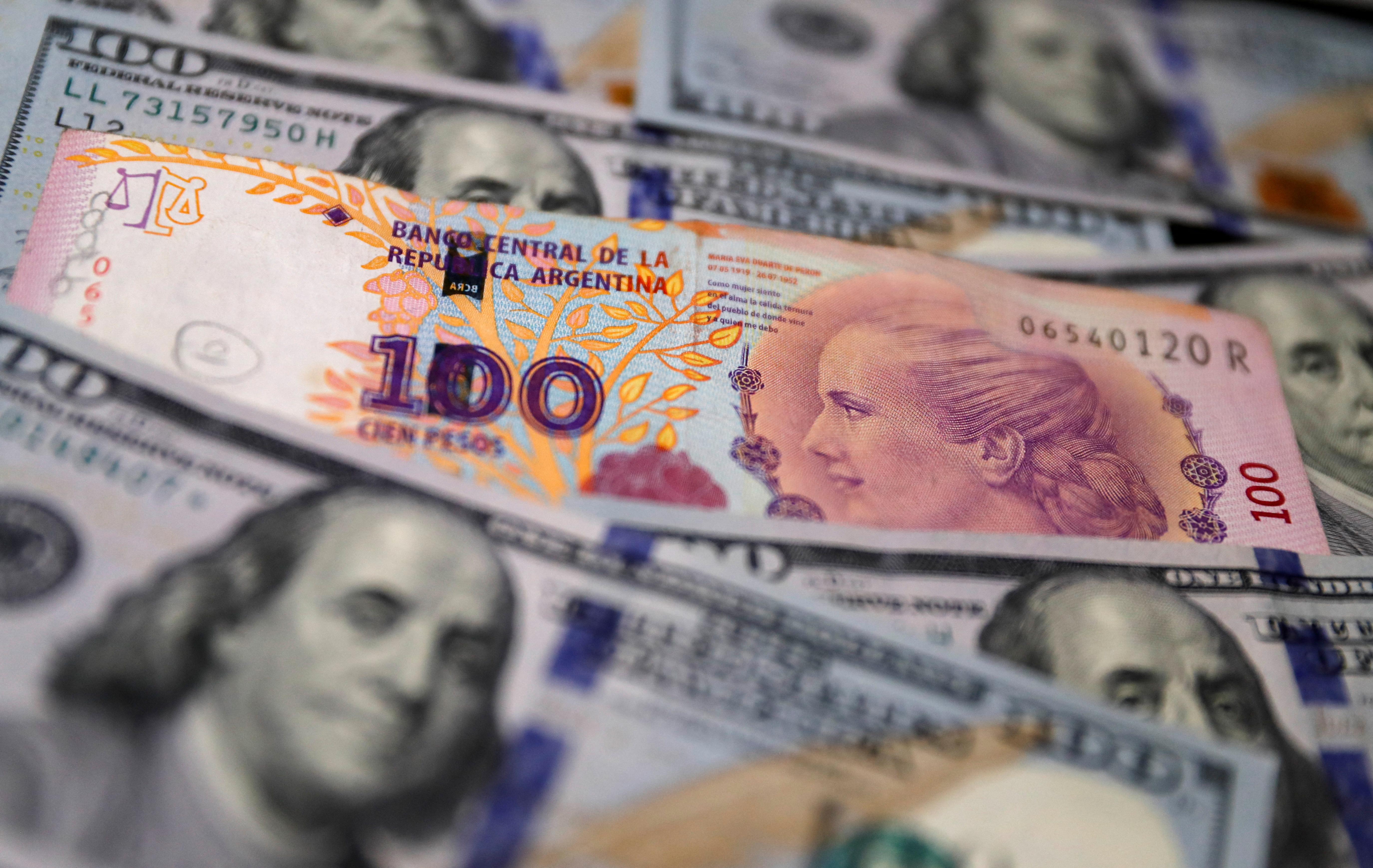 ورقة نقدية من فئة مائة بيزو أرجنتيني تجلس فوق عدة أوراق نقدية من فئة مائة دولار أمريكي في هذه الصورة التوضيحية